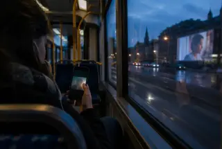Passenger watching a vertically framed film on a smartphone while a cinema screen reflects in a tram window.